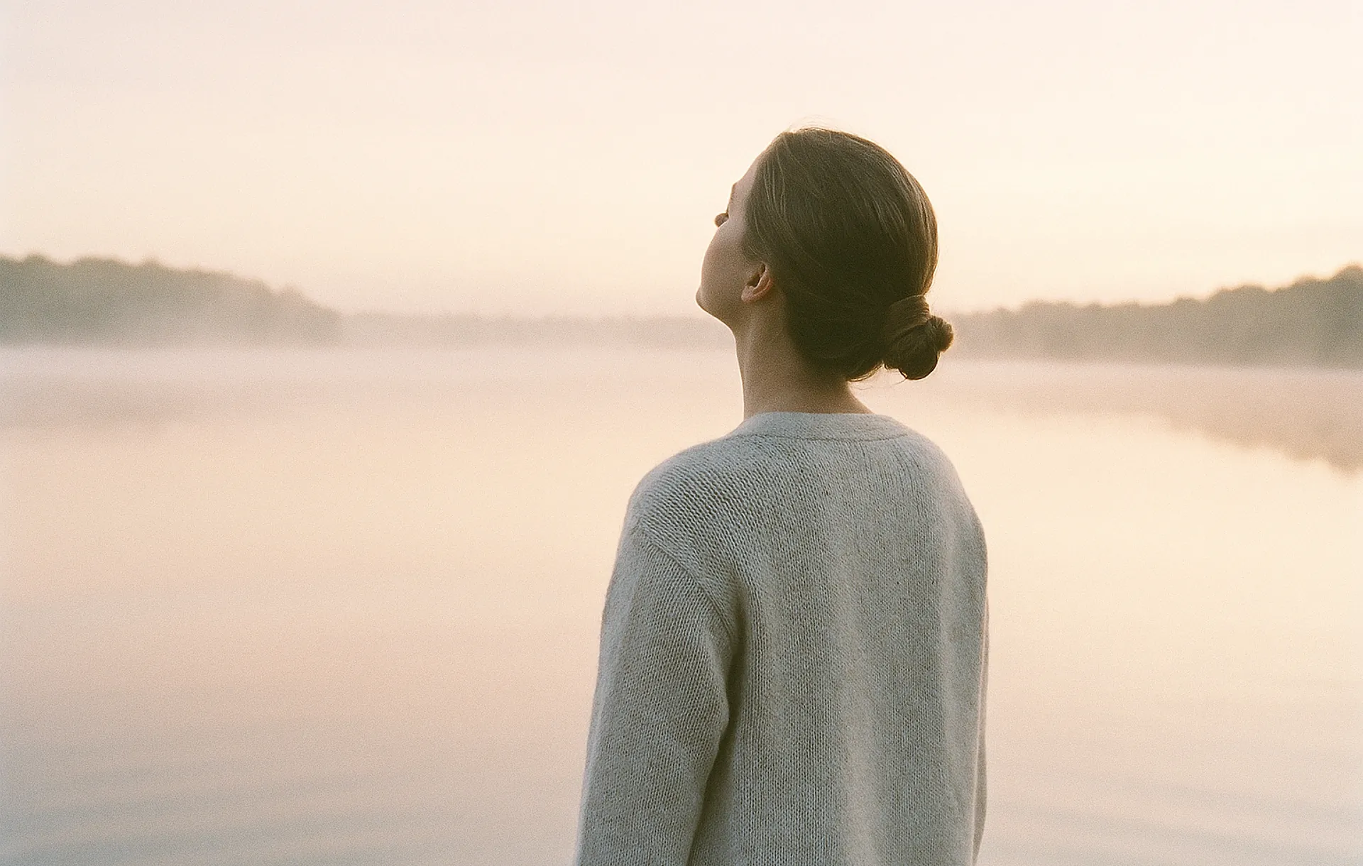 Une femme de dos respire profondément face à un lac calme au lever du soleil, brume légère